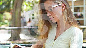 Woman in eyeglasses reading book sitting at table in outdoor cafe