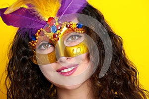 Woman face closeup in carnival masquerade mask with feather, beautiful girl portrait on yellow color background, long curly hair