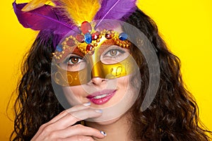 Woman face closeup in carnival masquerade mask with feather, beautiful girl portrait on yellow color background, long curly hair