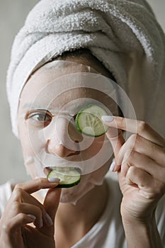 A woman in a face mask is having fun with round cucumbers, looking at the camera and biting a cucumber. Natural