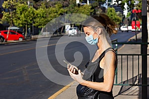 Woman with face mask on looking at cellphone while waiting on bus stop