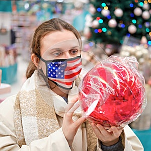 Woman in face mask with usa flag buys christmas baubles in gift shop. Lifestyle, a celebration of the coronavirus pandemic