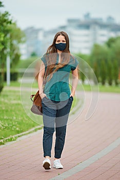 Woman in a face mask walks thrusting hands into pockets of trousers in the park