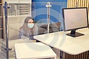 A woman in a face mask works on a computer in a room with glass partitions. Working with virus protection during an influenza
