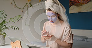 Woman with facial mask and towel on her head working with smartphone at home