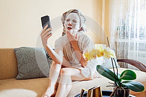 Woman with facial sheet mask applied wearing towel on head relaxing at home after bath using smartphone