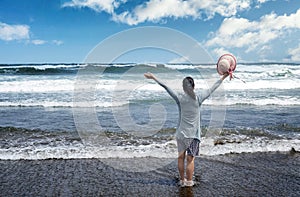 Woman facing ocean and sky with arms up while holding her hat