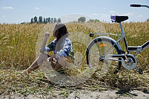 Woman farmer sitting on a wheat field. Young pretty farmer woman on farmland with bicycle. Beautiful girl in plaid shirt in golden