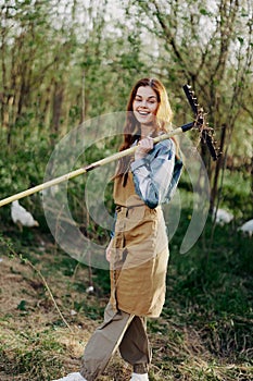 A woman farmer in work clothes and an apron works outdoors in nature and holds a rake to gather grass