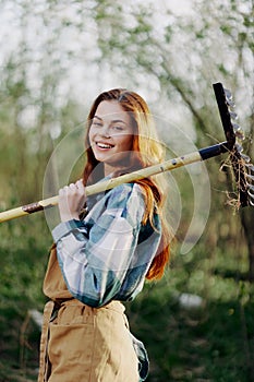 A woman farmer in work clothes and an apron works outdoors in nature and holds a rake to gather grass