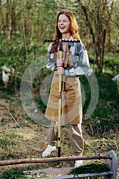 A woman farmer in work clothes and an apron works outdoors in nature and holds a rake to gather grass