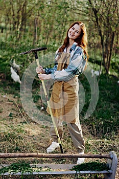 A woman farmer in work clothes and an apron works outdoors in nature and holds a rake to gather grass