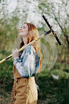 A woman farmer in work clothes and an apron works outdoors in nature and holds a rake to gather grass