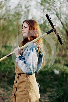 A woman farmer in work clothes and an apron works outdoors in nature and holds a rake to gather grass