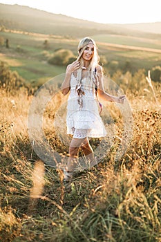 Woman fashion model portrait outdoors in the summer field, sunset. Boho style young woman in white dress and with