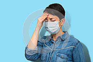 Woman feeling strong headache holding head on light blue background. Middle aged brunette in medical mask frown in pain