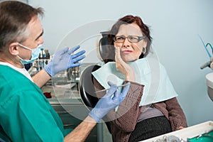 Woman feeling toothache, touching cheek with hand at dental clinic. Senior dentist trying to help