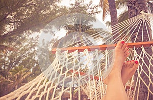 Woman Feet in a hammock with palm trees and bright sunset. Vacation concept with orange color tone