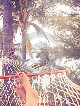 Woman Feet in a hammock with palm trees and bright sunset. Vacation concept with orange color tone