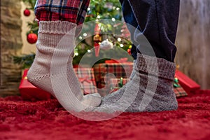 Woman feet standing in tip toe in winter socks on male lags on a fluffy red blanket near a Christmas tree with gifts. Concept.