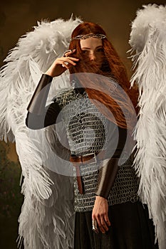 Woman with fiery red hair and large white wings dressed medieval armor against vintage studio background. Warrior angel