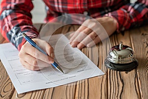 Woman filling hotel reservation form on wooden rustic reception desk with silver vintage bell. Hotel service, registration.