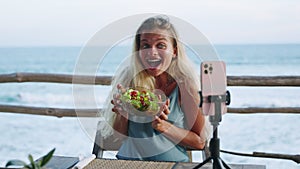 Woman films video of herself about lunch on tropical island. Female blogger holds bolw of green healthy salad and smiles