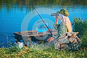 Woman fisherman carefully looks through binoculars on the river bank. Summer, hobby, vacation.