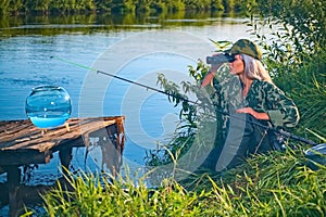 Woman fisherman carefully looks through binoculars on the river bank. Summer, hobby, vacation.