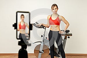 Woman fitness instructor using phone camera on tripod for online workout on exercise bike.