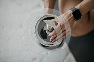 Woman with fitness tracker sits on floor and adjusts weight of dumbbell for hand exercises at gym