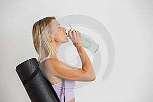 Woman fitness trainer holding yoga mat and drinking water from refillable bottle. Motivation concept