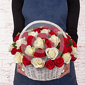 Woman florist in blue apron holding fresh bouquet of red and white flowers in basket against light brick wall background