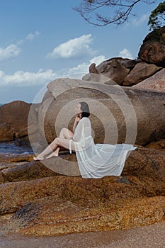 Woman in a flowing white dress enjoys a peaceful moment on coastal rocks, the sea softly lapping near her