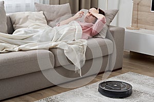 Woman with flu lying on sofa while robot vacuum cleaner is cleaning carpet in living room