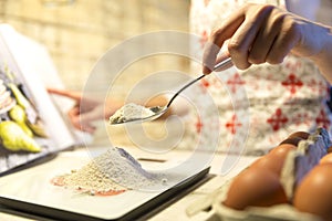 Woman following recipe in cookbook and measuring flour on kitchen scale