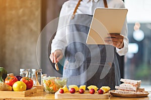 A woman following recipe on digital tablet while cooking salad and sandwich in the kitchen, online learning cooking class