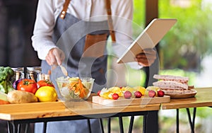 A woman following recipe on digital tablet while cooking salad and sandwich in the kitchen, online learning cooking class