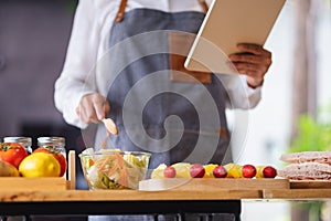 A woman following recipe on digital tablet while cooking salad and sandwich in the kitchen, online learning cooking class