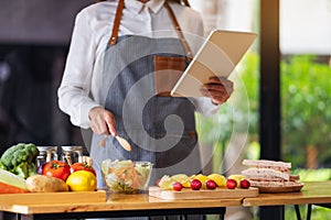 A woman following recipe on digital tablet while cooking salad and sandwich in the kitchen, online learning cooking class