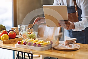 A woman following recipe on digital tablet while cooking salad and sandwich in the kitchen, online learning cooking class