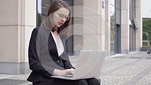 Woman in formal attire using laptop outside office building