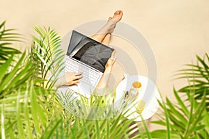 Woman freelancer with laptop sitting under palm tree branches. Sunscreen, sunglasses, orange juice on the table of sandy beach