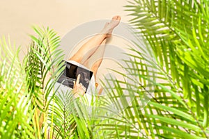 Woman freelancer in straw hat sitting under palm tree  branches on the sand of beach and working on tablet. Summer vacation.