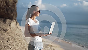 Woman freelancer in sunglasses holds laptop, looking on ocean at tropical beach