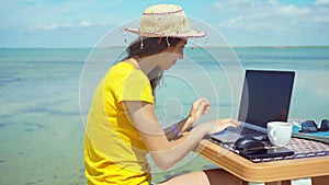 woman freelancer working on laptop computer, keyboarding text and holding cup with coffee on beach.