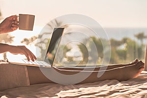 Woman freelancer working with laptop holding cup of coffee sitting on the bed in bedroom near panoramic window with view on palm