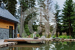 Woman, freelancer working on laptop, sitting on a pier by the backyard lake, a concept of remote office, work during