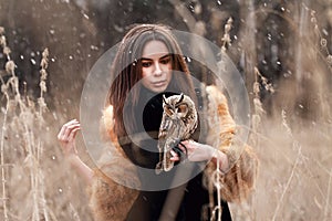 Woman in fur coat with owl on hand by first autumn snow. Beautiful brunette with long hair in nature, holding an owl. Romantic, d