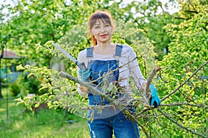 Woman gardener with saw and dry cut branches looking at camera in garden
